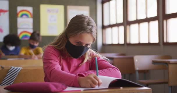 学校の机の上に座っている間 顔のマスクを身に着けて 本に書いている白人の少女 Covid19時の初等教育社会的距離の健康上の安全性スローモーションでコロナウイルスパンデミック — ストック動画