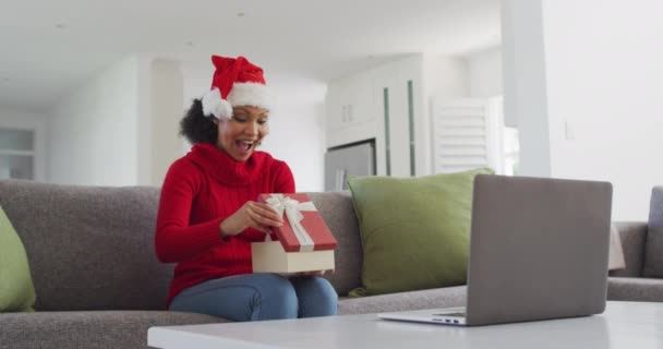 自宅のリビングルームのソファに座っているアフリカ系アメリカ人の女性は クリスマスプレゼントを開き 帽子をかぶっています 家で一人で過ごす女 — ストック動画