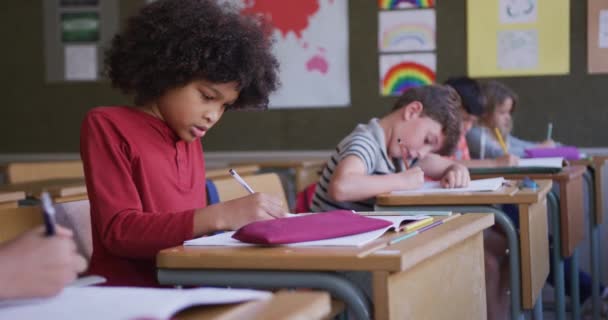 学校の机の上に座っている間に本に書いている複雑な人種の少女 背景の子供たち Covid19時の初等教育社会的距離の健康上の安全性スローモーションでコロナウイルスパンデミック — ストック動画