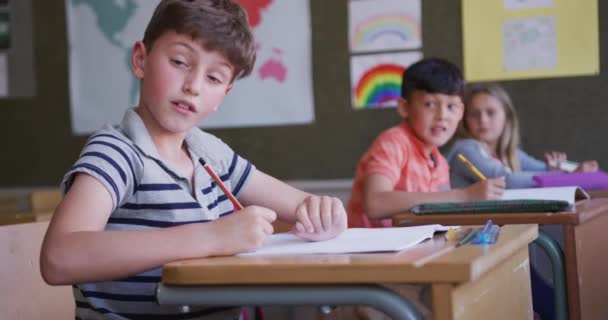学校の机の上に座りながら本を書いている白人の少年は微笑んだ Covid19時の初等教育社会的距離の健康上の安全性スローモーションでコロナウイルスパンデミック — ストック動画