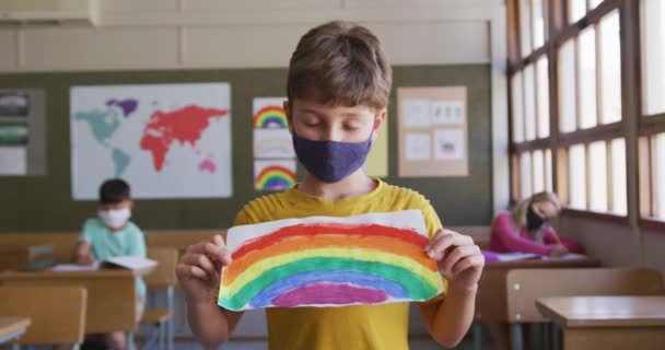 学校の教室で笑顔で虹の絵を描いたフェイスマスクをした白人少年 Covid19時の初等教育社会的距離の健康上の安全性スローモーションでコロナウイルスパンデミック — ストック動画