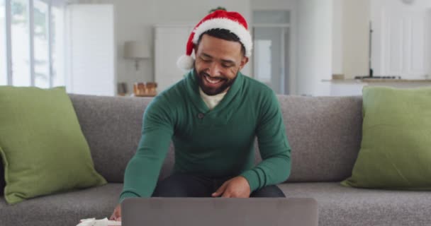 自宅のリビングルームでソファに座っているアフリカ系アメリカ人の男性は 帽子をかぶって クリスマスプレゼントを開きます 家で一人で過ごす男 — ストック動画