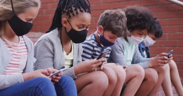 Grupa Wieloetnicznych Dzieciaków Noszących Maski Twarzy Pomocą Smartfonów Siedzących Ścianie — Wideo stockowe