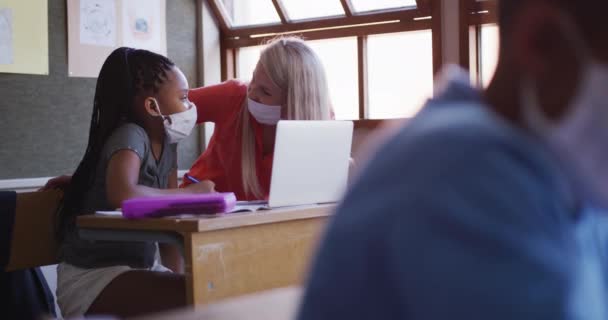 混合种族女孩在学校使用她的笔记本电脑 她的老师帮助她 所有的人都戴着口罩 Covid19 Coronavirus流感大流行期间 初等教育在社会上与健康安全保持距离的慢动作 — 图库视频影像