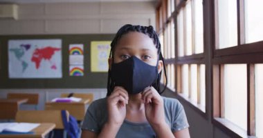 Yüz maskesi takan melez bir kız, okulda masasında oturuyor, kameraya bakıyor. Covid19 Coronavirus salgını sırasında temel eğitim sosyal güvenlikten uzaklaşma yavaş çekimde.