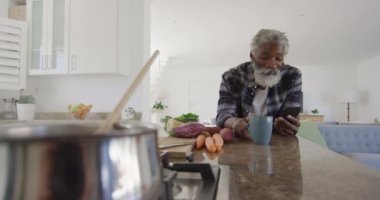 Afrika kökenli Amerikalı kıdemli bir adam mutfakta duruyor, akıllı telefon kullanıyor, sosyal uzaklık ve karantina tecridinde kendi başına kalma, yavaş çekim.
