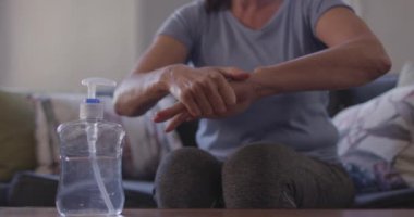 Beyaz kadın evde vakit geçiriyor, sosyal uzaklık ve tecrit karantinada, oturma odasında kanepede oturuyor, ellerini dezenfekte ediyor Covid 19 Coronavirüs 'ten koruyor, yavaş çekim.