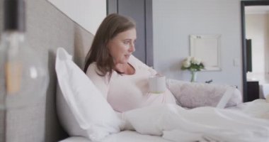 Mutlu beyaz kadın evde vakit geçiriyor, sosyal mesafeler ve karantinada kendini izole ediyor, yatak odasında uzanıyor, çay içiyor, dizüstü bilgisayarını yavaş çekimde kullanıyor..