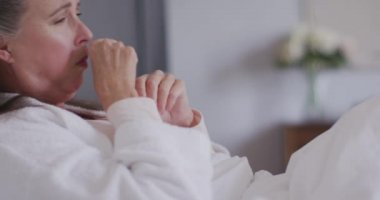 Hasta Kafkasyalı kadın evde vakit geçiriyor, sosyal uzaklık ve kendini karantinaya almış, yatakta uzanıyor, elinde doku tutuyor ve ağır çekimde öksürüyor..