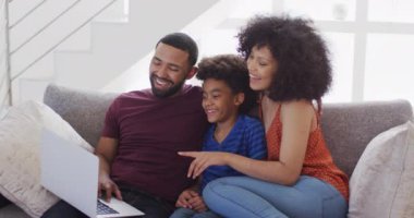 Karışık ırk çifti ve oğlu evde birlikte vakit geçiriyorlar. Sosyal mesafeler ve karantina tecridi, kanepede oturup dizüstü bilgisayar kullanıyorlar ve gülümsüyorlar, yavaş çekim.