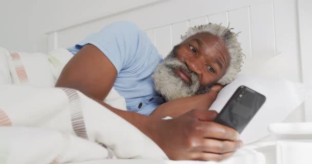 Старший Афроамериканец Лежит Кровати Спальне Используя Смартфон Улыбаясь Социальное Дистанцирование — стоковое видео