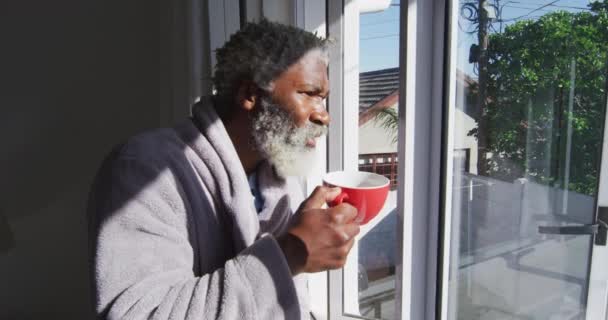 非裔美国老人站在卧室里 举着一杯咖啡 从窗户往外看 与外界保持距离 在隔离中自我隔离 行动缓慢 — 图库视频影像