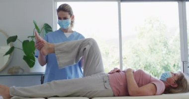 Kafkas kadın hemşire tarafından ziyaret edilmiş, bacağını esnetiyor, maske takan hemşire. Covid 19 Coronavirus karantinası sırasında evde tıbbi bakım, yavaş çekim