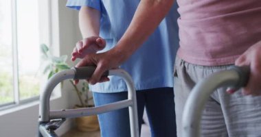 Kafkas kadın hemşire tarafından ziyaret edilen, kıdemli Kafkasyalı kadın, bir yürüteç kullanarak yürüyor, maske takan hemşire. Covid 19 Coronavirus karantinası sırasında evde tıbbi bakım, yavaş çekim