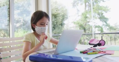 Beyaz kız evde maske takarak, ödevlerini yaparken ağır çekimde dizüstü bilgisayar kullanarak vakit geçiriyor. Covid 19 Coronavirus karantina süresince sosyal mesafeler.