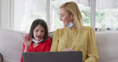Beyaz bir kadın kızıyla evde vakit geçiriyor. Yüz maskeleri takıyor, laptopta yavaş çekimde görüntülü sohbet ediyor. Covid 19 Coronavirus karantina süresince sosyal mesafeler.