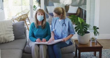 Kafkas kadın hemşire tarafından ziyaret edilen, evde kıdemli beyaz kadın, elleri kullanarak kitap okuyan, maske takan hemşire. Covid 19 Coronavirus karantinası sırasında evde tıbbi bakım, yavaş çekim.