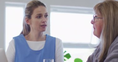 Senior Caucasian woman at home visited by Caucasian female nurse, sitting by table and talking. Medical care at home during Covid 19 Coronavirus quarantine, slow motion.