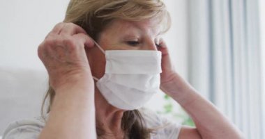 Üst düzey beyaz kadın evde oturuyor, yatağında oturuyor, maske takıyor. Covid 19 Coronavirus karantina süresince sosyal mesafeler