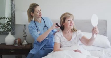 Kafkas kadın hemşire tarafından ziyaret edilen, saçlarını tarayan, ayna kullanan, kıdemli beyaz kadın. Covid 19 Coronavirus karantinası sırasında evde tıbbi bakım, yavaş çekim.