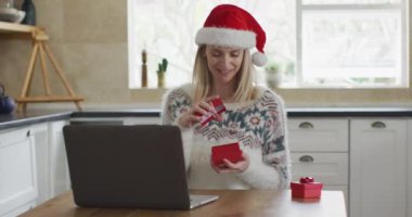Beyaz kadın evde vakit geçiriyor, Noel 'de mutfakta oturuyor, Noel Baba şapkası takıyor, dizüstü bilgisayar kullanıyor ve hediyeyi açıyor. Covid 19 Coronavirus karantinası sırasındaki sosyal mesafeler, yavaş çekim.