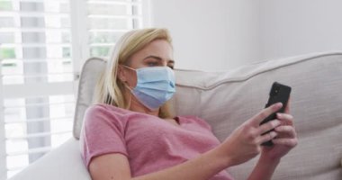 Beyaz kadın evde maske takarak, akıllı telefonu yavaş çekimde koltukta yatarak geçiriyor. Covid 19 Coronavirus karantina süresince sosyal mesafeler.