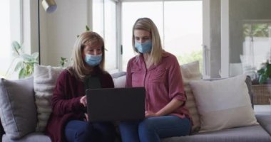 Senior Caucasian woman spending time at home with her adult daughter, sitting on couch talking and using laptop computer. Social distancing during Covid 19 Coronavirus quarantine, slow motion