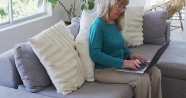 Üst düzey beyaz kadın evinde oturuyor, oturma odasında dizüstü bilgisayar kullanıyor. Covid 19 Coronavirus karantina süresince sosyal mesafeler