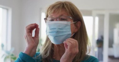 Üst düzey beyaz kadın, oturma odasında maske takarak vakit geçiriyor. Covid 19 Coronavirus karantina süresince sosyal mesafeler