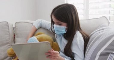 Beyaz kız evde vakit geçiriyor, oyuncak ayısına sarılıyor yüz maskeleri takıyor ve dijital tablet kullanıyor, ağır çekimde. Covid 19 Coronavirus karantina süresince sosyal mesafeler.