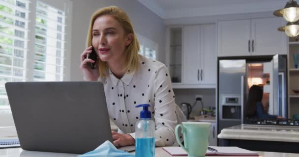 Fehér Otthon Tölti Idejét Maszkban Okostelefonon Beszél Laptopot Használ Konyhában — Stock videók