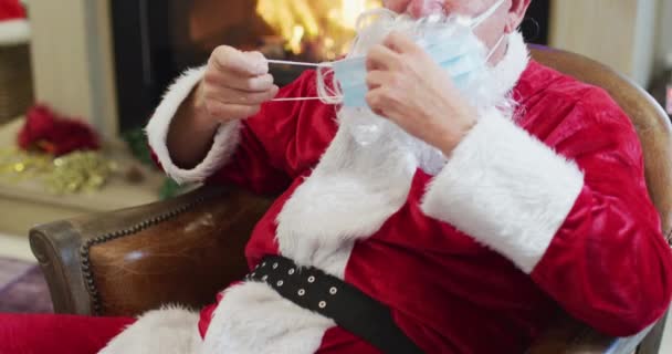 サンタクロースの肖像画は ゆっくりとした動きで クリスマスの間に自宅で暖炉のそばの椅子に座って顔のマスクを置く Covid 19コロナウイルス隔離ロックダウン中の社会的距離 — ストック動画