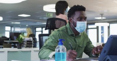 Sıradan bir ofiste çalışan Afrika kökenli Amerikalı bir adam, yüz maskesi takmış, Covid 19 'dan koruyor bilgisayarlı ağır çekimde masada oturuyor. Coronavirus Covid 19 salgını sırasında işyerinde sosyal mesafeler.