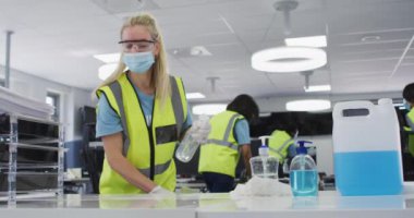 Ofisi temizleyen beyaz kadın çelik yelek, eldiven ve yüz maskesi takıyor, dezenfektan kullanıyor, arka planda iş arkadaşları, ağır çekim. Coronavirus Covid 19 salgını sırasında işyerinde hijyen