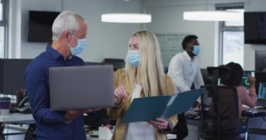 Kafkas erkek ve kadın günlük ofiste çalışıyor, maske takıyor ayakta durmaktan koruyor, dizüstü bilgisayarı ağır çekimde tutuyor. Coronavirus Covid 19 salgını sırasında işyerinde sosyal mesafeler.