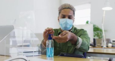 Ofiste maske takmış, el dezenfektanı ile el dezenfekte eden melez erkek yaratıcı ağır çekimde oturuyor. Coronavirus Covid 19 salgını sırasında işyerinde sağlık ve hijyen.