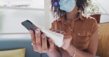 Ofisteki kanepede oturan, yüz maskesi takan akıllı telefonunu yavaş çekimde dezenfekte eden melez bir iş kadını. Coronavirus Covid 19 salgını sırasında işyerinde sağlık ve hijyen.