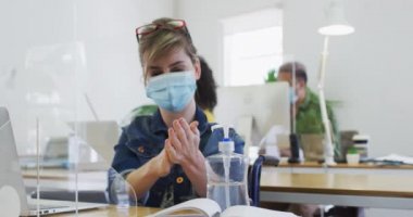 Beyaz kadın ofisinde koruyucu ekranlı, maske takmış, ağır çekimde ellerini dezenfekte ediyor. Coronavirus Covid 19 salgını sırasında işyerinde sağlık ve hijyen.