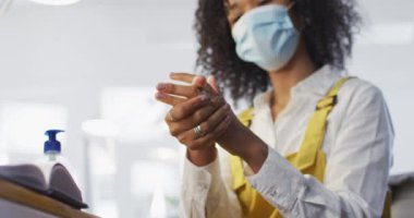 Ofiste maske takmış, el dezenfektanı ile el dezenfekte eden melez, yaratıcı kadın. Coronavirus Covid 19 salgını sırasında işyerinde sağlık ve hijyen.