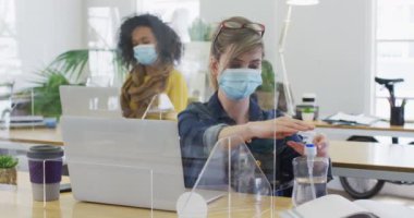 Beyaz kadın ofisinde koruyucu ekranlı, maske takmış, ağır çekimde ellerini dezenfekte ediyor. Coronavirus Covid 19 salgını sırasında işyerinde sağlık ve hijyen.