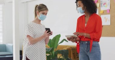 Karışık ırk ve Kafkas kadın iş adamları ofiste maske takarak, akıllı telefon ve tablet kullanarak, ağır çekimde konuşuyorlar. Coronavirus Covid 19 salgını sırasında işyerinde sağlık ve hijyen.