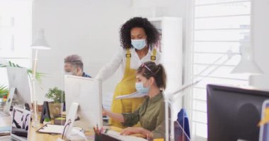 Karışık ırk ve Kafkas kadın yaratıcı iş arkadaşı yüz maskeleri takarak ofiste konuşuyor, arka planda erkek meslektaşı var. Coronavirus Covid 19 salgını sırasında işyerinde sağlık ve hijyen.