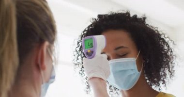 Beyaz kadın dijital termometre kullanarak işe yüz maskesi takarak gelen melez kadının ateşini ölçüyor. Coronavirus Covid 19 salgını sırasında işyerinde sağlık ve hijyen.