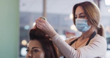 Kafkas kadın kuaför yüz maskesi takarak kuaförde çalışıyor, Kafkas kadın saçlarına bigudi takıyor, ağır çekimde. Coronavirus Covid 19 salgını sırasında işyerinde sağlık ve hijyen.