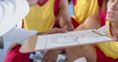 Afro-Amerikan basketbol koçu kırmızı formalı oyunculara bir panoda strateji sıraladı. Oyun diyagramına odaklan, bir spor bağlamında bir taktik planlama anını vurgula.