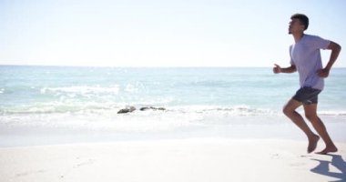 Çift ırklı genç bir adam fotokopi alanı ile güneşli bir plaj boyunca koşuyor, spor kıyafetleri ile vurgulanan atletik formu. Yavaş çekimde deniz kenarında çalışmaktan hoşlanıyor..