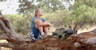 Genç beyaz kadın devrilmiş bir ağaçta oturuyor, düşünceli görünüyor. Doğa ile çevrili, yürüyüş sırasında dinleniyor, yanında bir sırt çantasıyla, ağır çekimde..