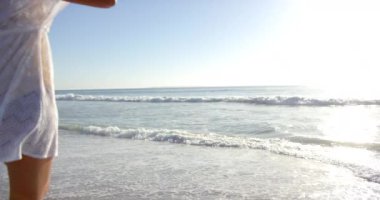 Genç bir melez kadın kıyı boyunca yürümekten zevk alıyor, beyaz elbisesi rüzgarda uçuşuyor. Onun rahat duruşu ve sakin plaj düzeni huzur ve rahatlık hissi uyandırıyor..