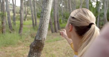 Sarı saçlı genç bir kadın kameraya gülümsüyor ve kolunu fotokopi aletiyle ağaçlara doğru uzatıyor. Dışarıda güneşli bir orman ortamında, yavaş çekimde bir neşe anı yakalıyor..