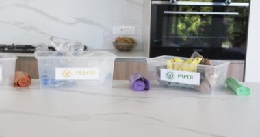 Cam, plastik ve kağıt ayırmak için etiketlenmiş üç kutu fotokopi alanı olan bir mutfak tezgahında. Çöp kutuları, şişeler ve meyve kabukları gibi görünen içeriklerle modern bir mutfak zeminine karşı geri dönüşümü teşvik ediyor..